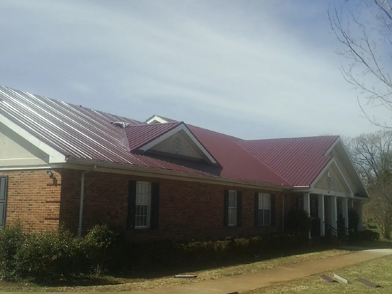 Red metal roof with gabled entries on a long brick building, from a Hail Damage Roof Repair job in West Hyannisport