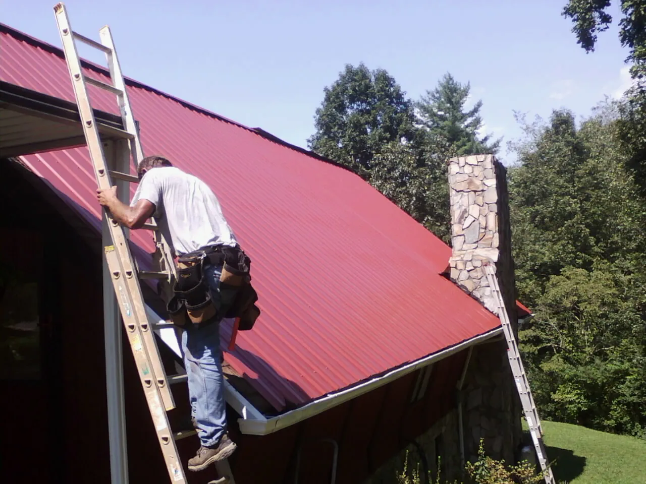 Roofer on a ladder beside a red metal roof and stone chimney — Soffit & Fascia Repair by Roofing Brothers in West Hyannisport
